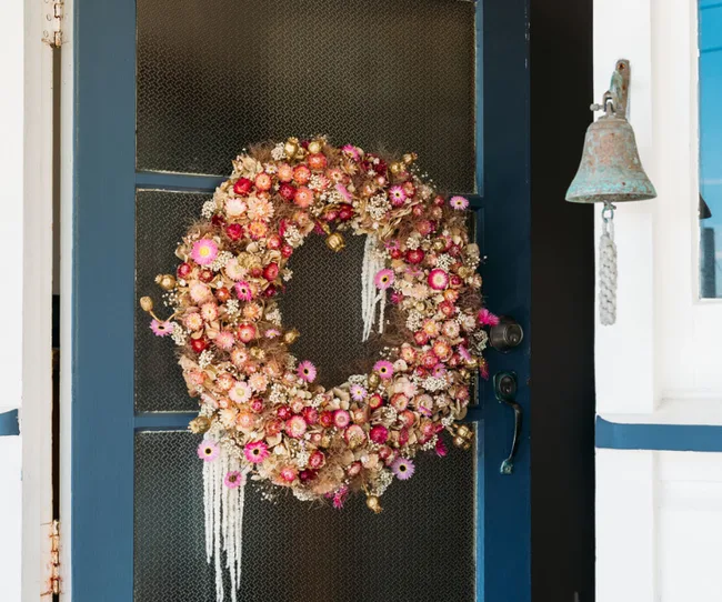 A vibrant dried flower wreath with pink and gold hues hangs on a blue door next to a rustic metal bell.
