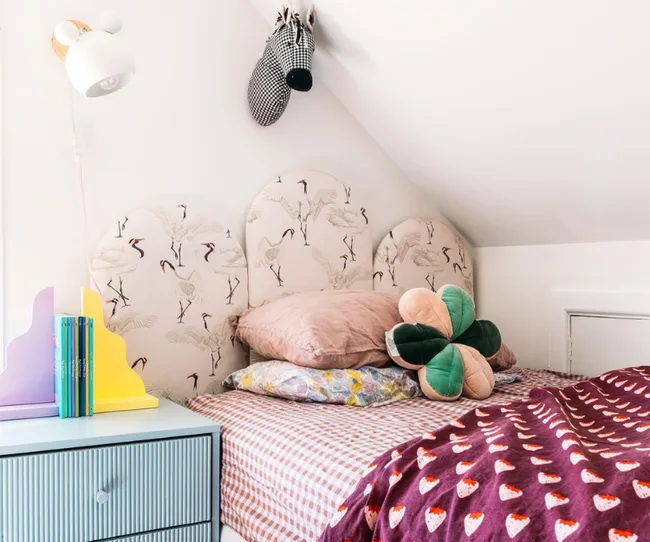 Cozy bedroom with patterned pillows, bird-themed headboard, wall-mounted lamp, and a colorful chest with books.