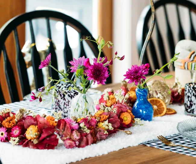 A dining table centerpiece with vibrant flowers in vases and dried oranges on a checkered tablecloth.