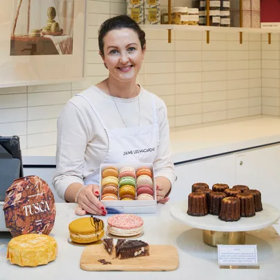 Woman in a bakery displaying a box of colorful macarons, with various pastries on the counter.