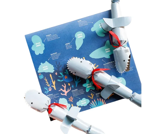 Three shark-shaped holiday crackers on a blue ocean-themed activity sheet with coral illustrations.