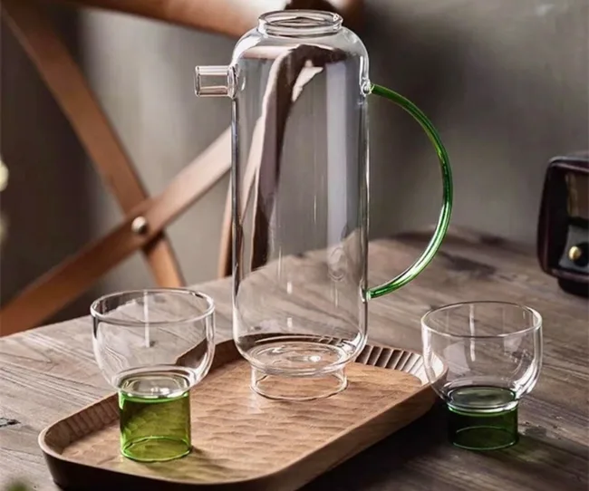 Glass jug with green handle and spout, on a wooden tray with two matching glasses, placed on a wooden table.