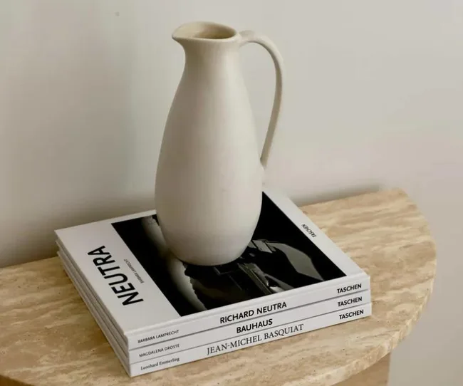 Cream-colored ceramic pitcher on stacked books about Richard Neutra, Bauhaus, and Jean-Michel Basquiat on a beige table.