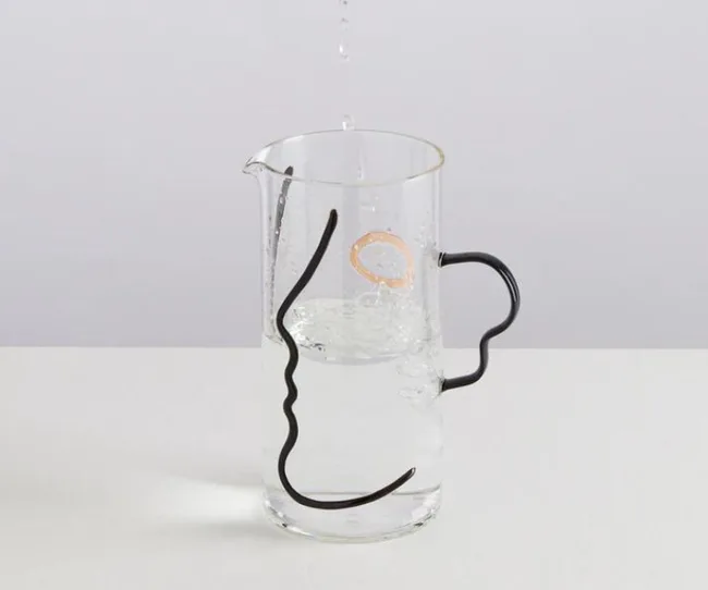 Glass pitcher with abstract line art of a face, half-filled with water, on a white surface.