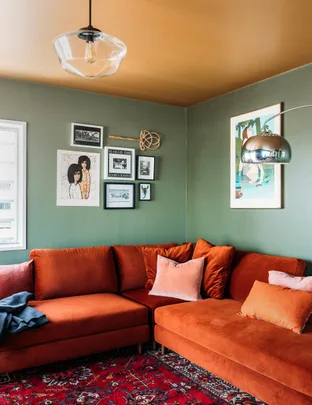 Cozy living room with orange velvet sofa, green walls, eclectic art, and decorative pillows on a patterned red rug.