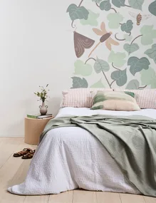 Bed with green blanket, plant-themed wallpaper, round bedside table, books, and vase in bright, stylish room.