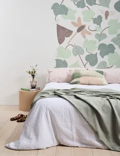 Bed with green blanket, plant-themed wallpaper, round bedside table, books, and vase in bright, stylish room.