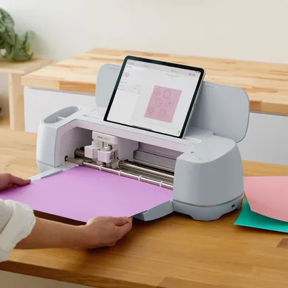 A person using a Cricut Maker 3 machine with a tablet displaying flower designs on a wooden table.
