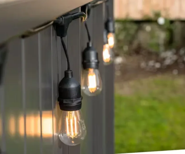 String lights with glowing bulbs hanging over a garden fence.