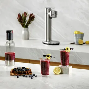 Two glasses of blueberry lemonade with a soda maker on a marble counter, garnished with lemon twists and berries.