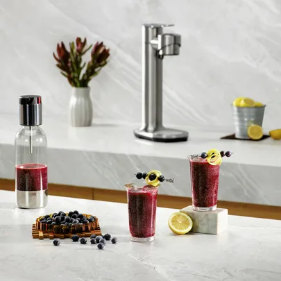 Two glasses of blueberry lemonade with a soda maker on a marble counter, garnished with lemon twists and berries.