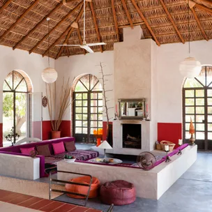 Tropical-themed living room with colorful cushions, thatched roof, and large windows surrounding a central fireplace.