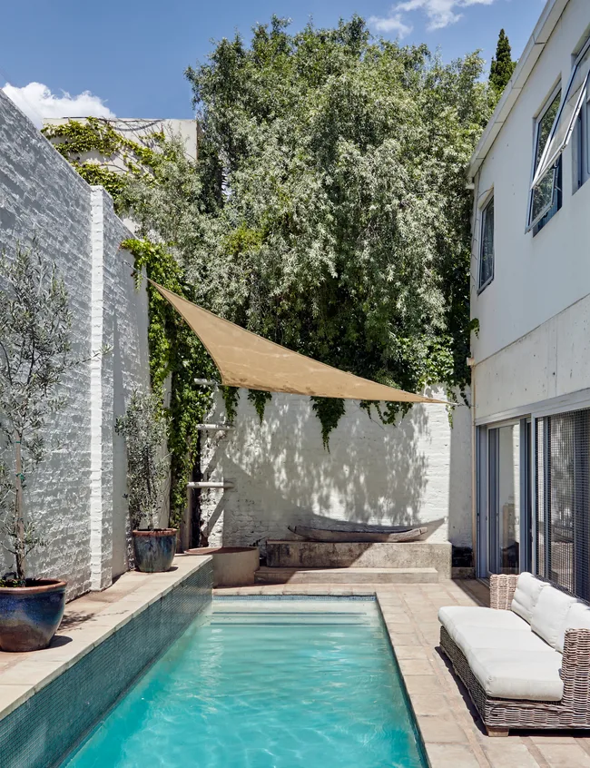 Outdoor pool with a wicker couch, white walls, potted plants, and a shade sail, adjacent to a modern building.