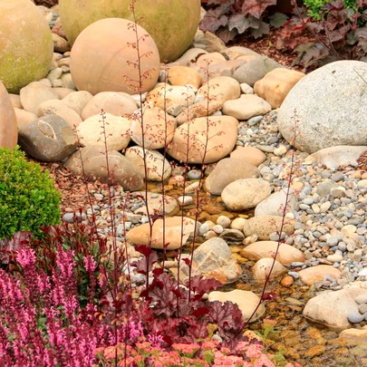Garden with round stones, small stream, and pink flowers in the foreground.