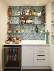 Bar area with shelves of liquor bottles, a mini-fridge stocked with cans, and tropical floral wallpaper background.
