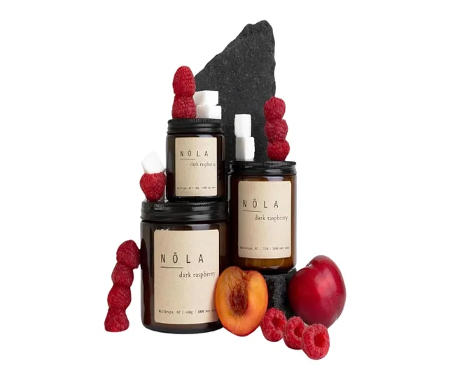 Brown jars labeled "NÖLA dark raspberry" surrounded by raspberries, a plum, and a slice of peach on a slate slab.