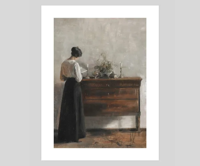 A woman in a white blouse and dark skirt stands at a wooden sideboard, reading a book.