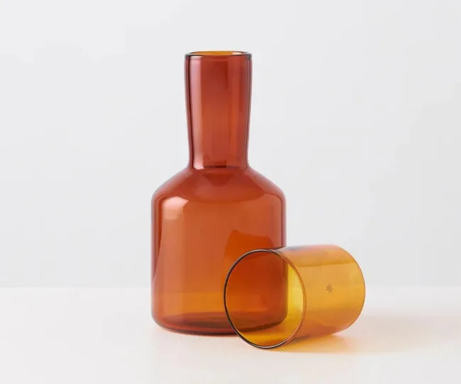 Amber glass carafe with a cup placed on its side, showcasing a minimalist and elegant design against a plain background.