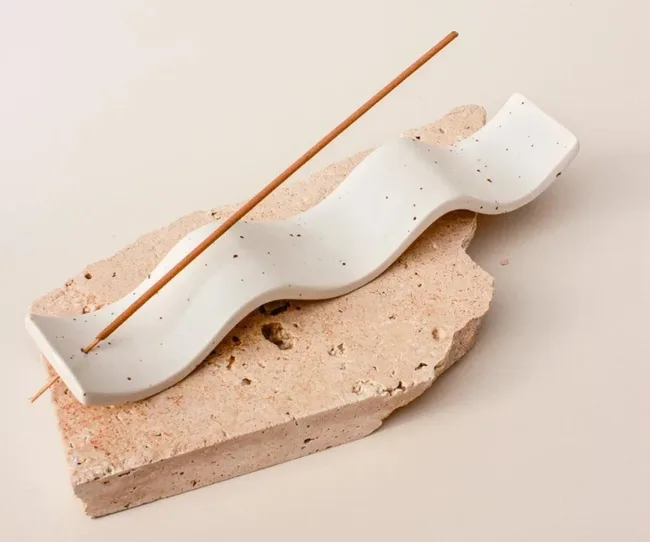 A wavy, speckled ceramic incense holder on a stone slab with a burning incense stick.