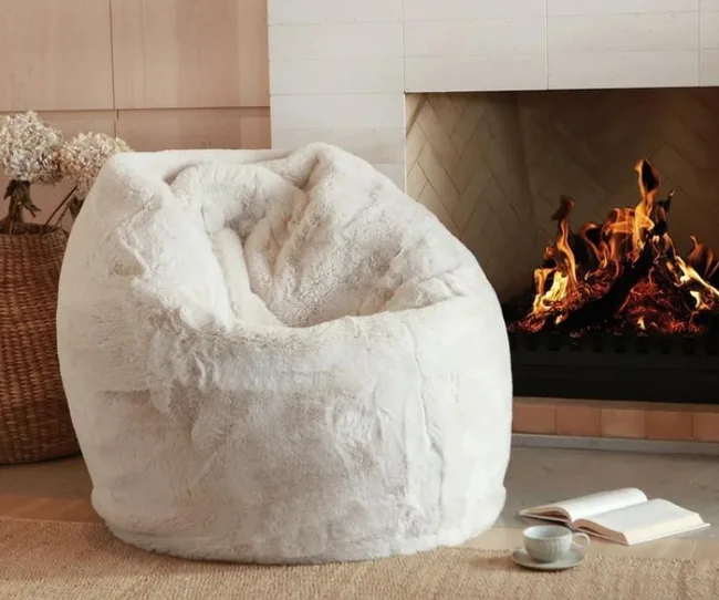 Cozy white bean bag in front of a lit fireplace, with an open book and steaming cup on the floor.