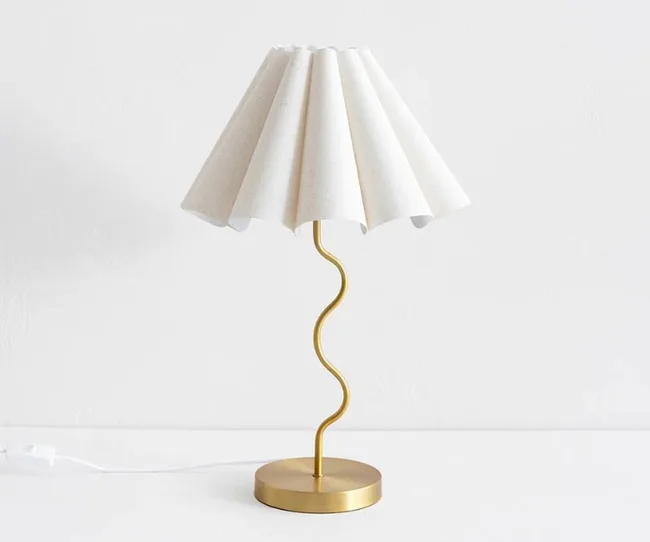 Gold lamp with a wavy stand and a pleated white shade, on a white surface against a light background.