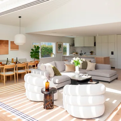Bright, modern living room with a gray sofa, white armchairs, dining table, and open kitchen.