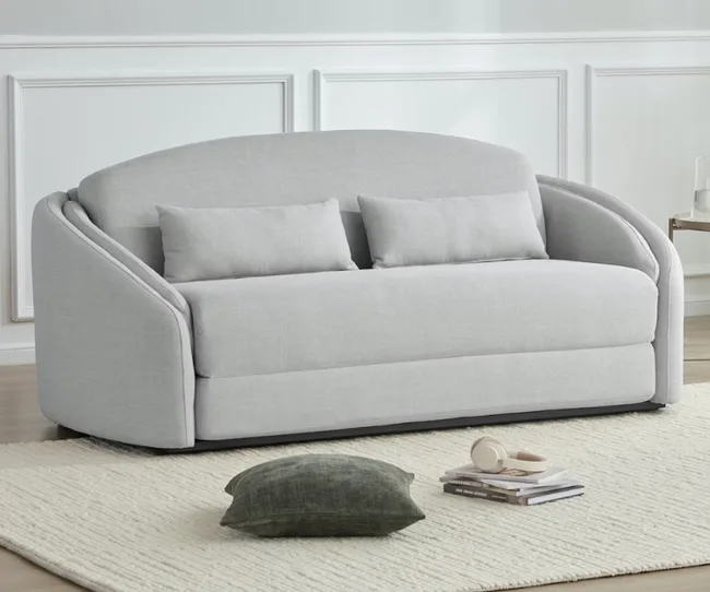 Light gray modern sofa with two cushions on a cream rug, accompanied by a small green pillow, books, and headphones.