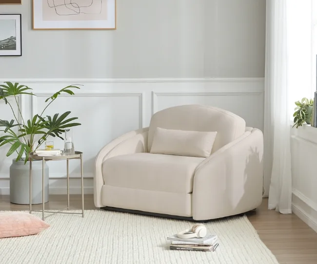Cream armchair in a bright room with a plant, side table, and books on a light rug.
