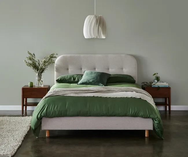 Modern bedroom with a beige headboard, green bedding, wooden nightstands, and a white pendant light.
