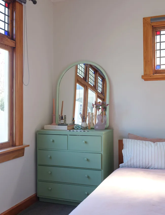 Bedroom corner with green dresser, mirror, books, and vases; bed with striped pillow and stained glass window reflections.
