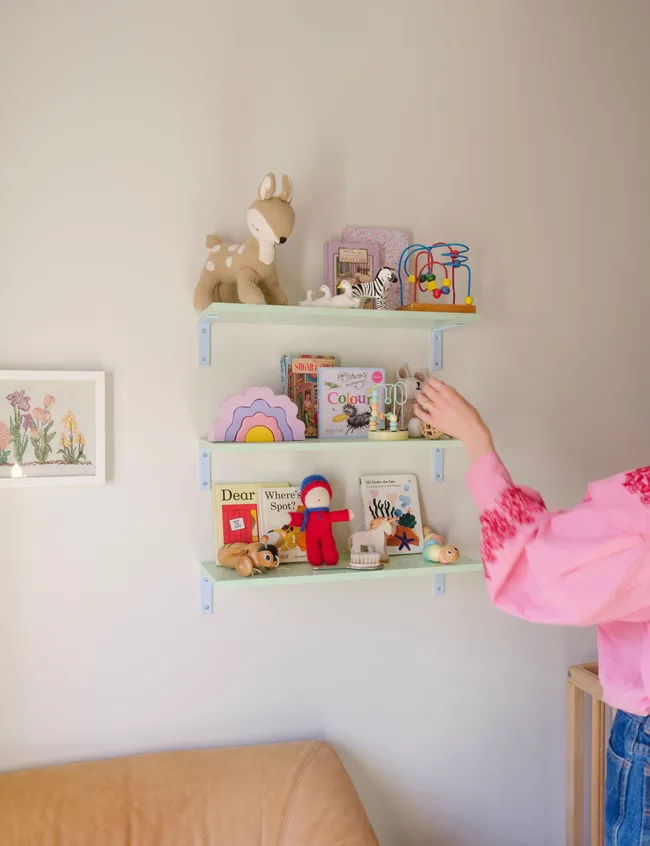"Wall shelves with children's toys and books; person in pink adjusts items."