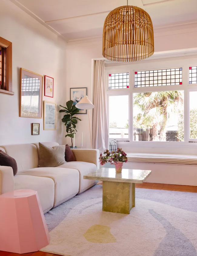 Cozy living room with a beige sofa, green marble coffee table, wall art, pink stool, large windows, and rattan pendant light.