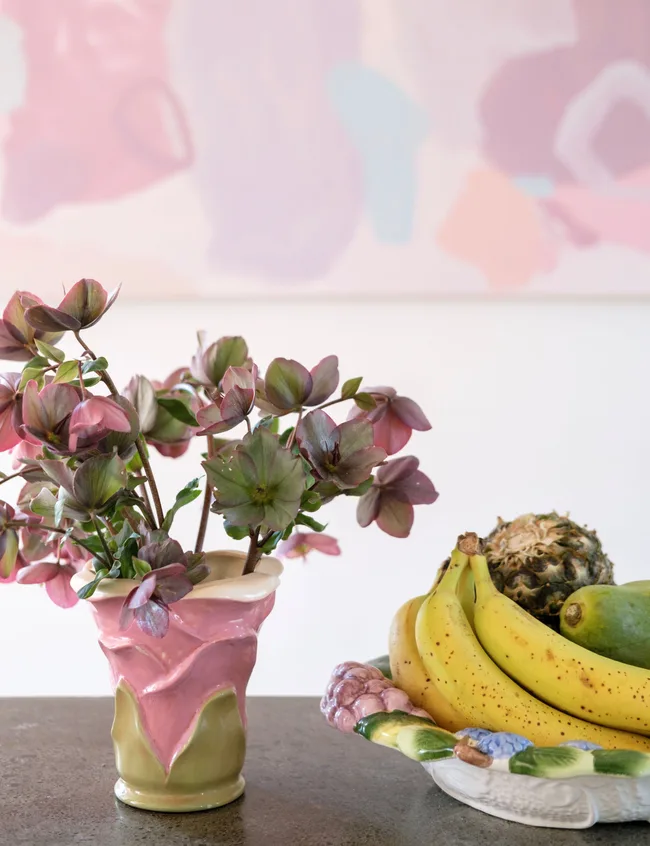 Vase with purple flowers and ceramic bowl with bananas, pineapple, pear; abstract painting in background.