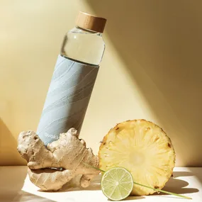 Glass bottle with a wooden cap, beside ginger root, pineapple slice, and a lime wedge on a light background.
