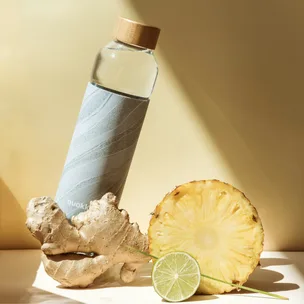 Glass bottle with a wooden cap, beside ginger root, pineapple slice, and a lime wedge on a light background.