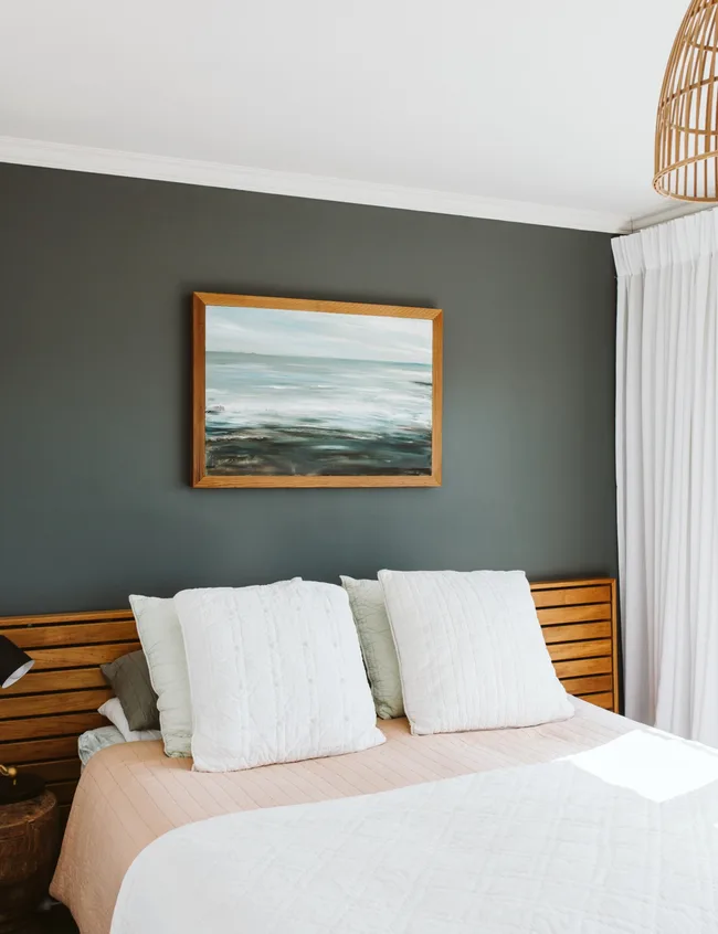 Bed with white pillows and beige quilt against dark wall, framed seascape painting above, wicker lamp on side.
