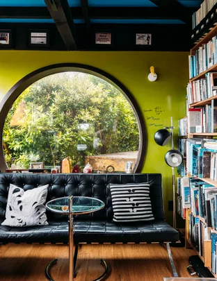 Cozy room with round window, black sofa, glass table, patterned cushions, and bookshelves filled with books.