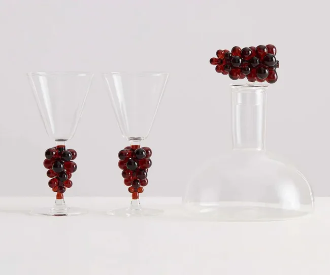Clear wine glasses and decanter with red grape-like embellishments on stems and stopper.