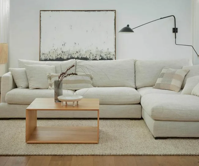 Contemporary living room with beige sectional sofa, modern wall lamp, abstract painting, and wooden coffee table.