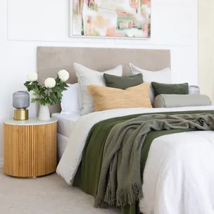 Cozy bedroom with green accents, pillows, and a wooden nightstand with flowers and a lamp.