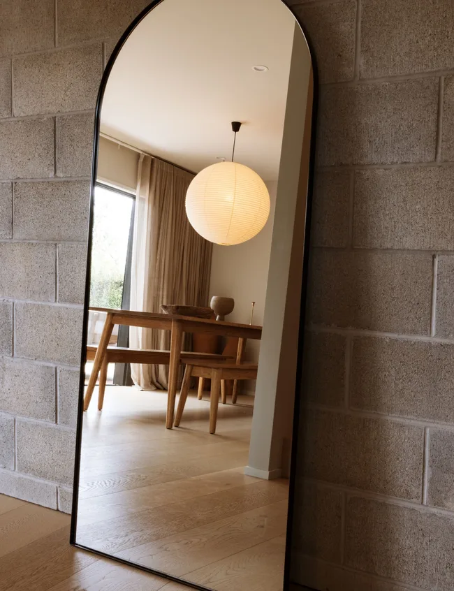 Reflection of a dining room with a wooden table and chairs in a tall, arched mirror against a brick wall.