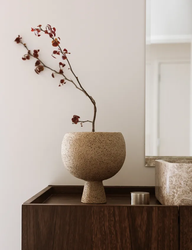 Ceramic vase with dried branch and red leaves on a wooden cabinet near a marble sink.