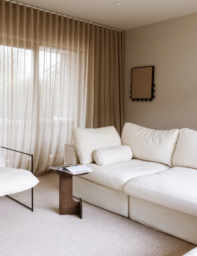 Cozy living room with beige sofa, sheer curtains, and a small wooden side table with an open magazine on it.