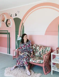A woman in a floral dress sits on a patterned sofa in a colorful room, holding a magazine and smiling.