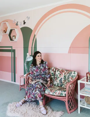 A woman in a floral dress sits on a patterned sofa in a colorful room, holding a magazine and smiling.