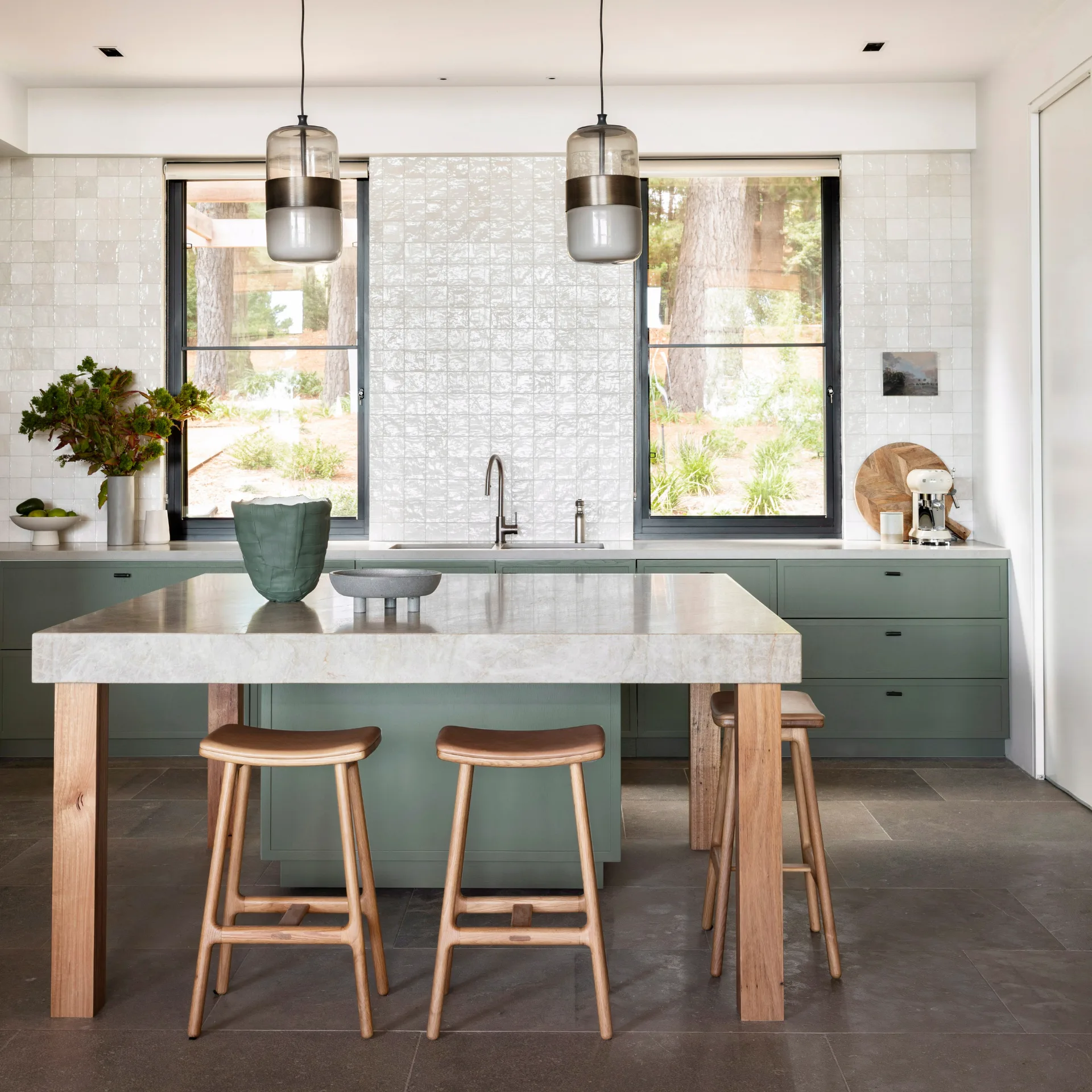 Modern kitchen with green cabinets, marble island, wooden stools, pendant lights, and large windows.