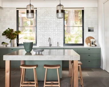 Modern kitchen with green cabinets, marble island, wooden stools, pendant lights, and large windows.