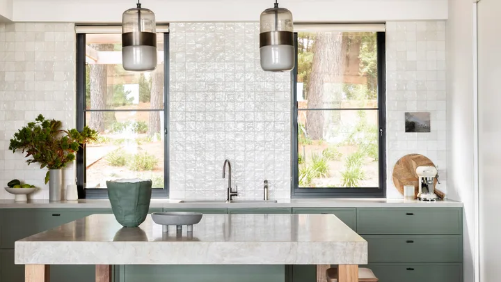 Modern kitchen with green cabinets, marble island, wooden stools, pendant lights, and large windows.