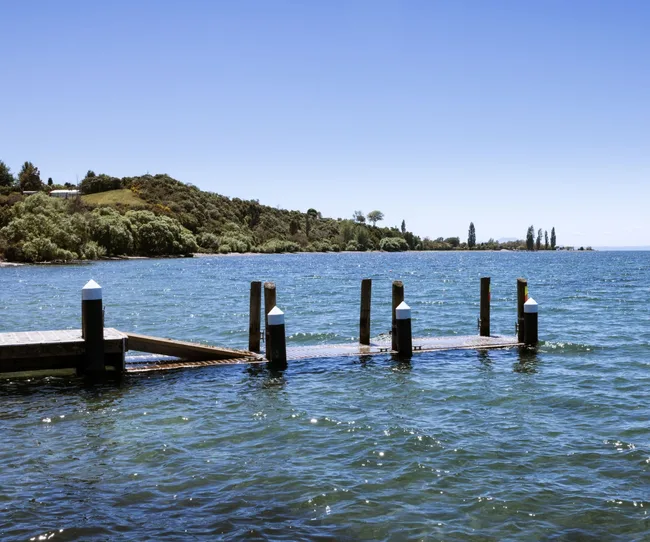 A wooden dock extends into a calm blue lake surrounded by green hills under a clear sky.