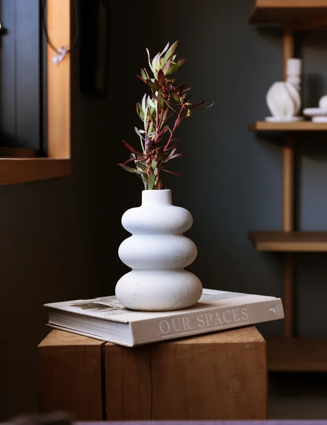 A white ceramic vase with flowers sits on a book titled "Our Spaces" on a wooden surface.